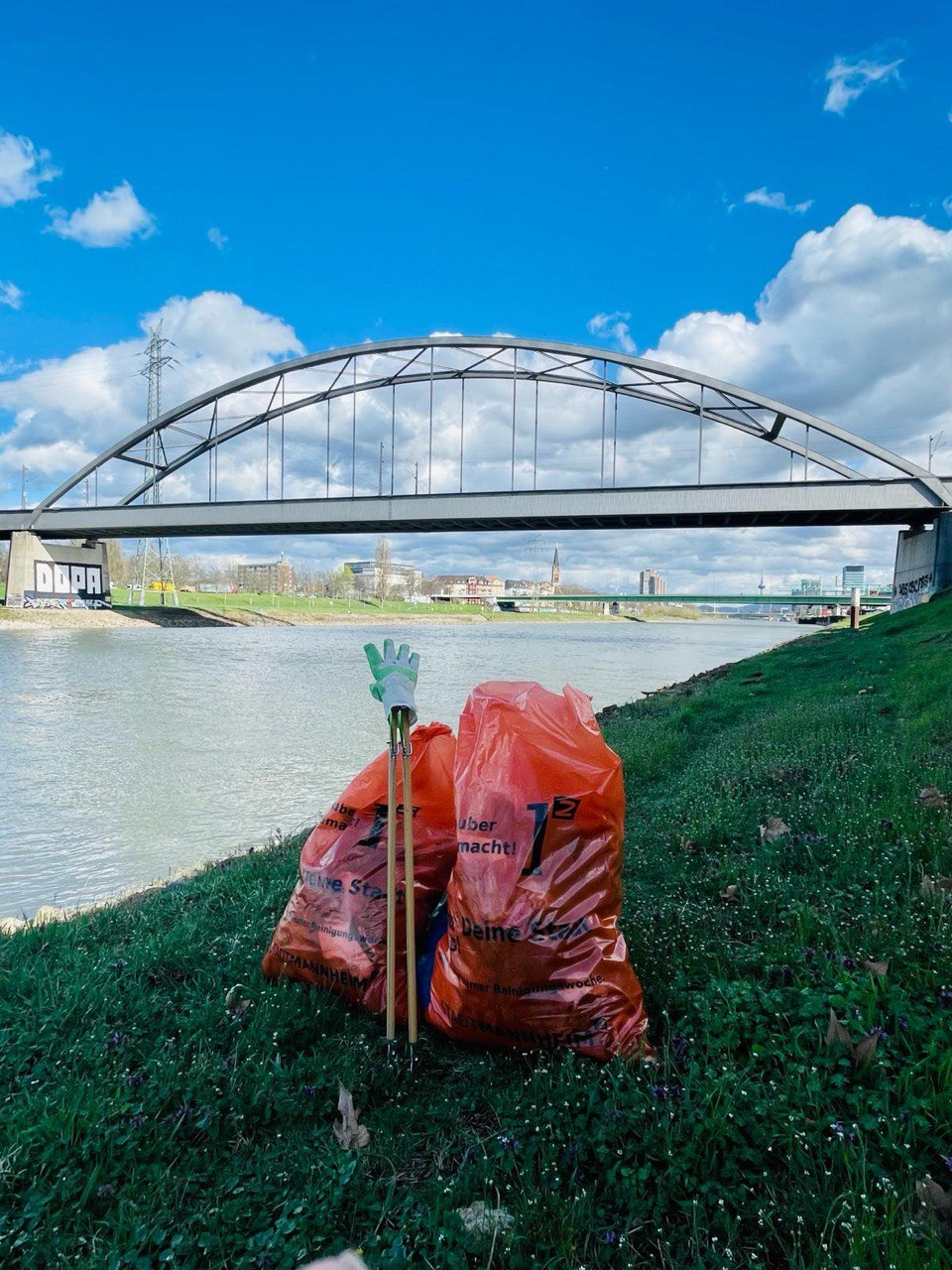 Man sieht die den Neckar und am Ufer zwei orangene Müllsäcke. Daneben stehen Müllpicker und Handschuhe. Im Hintergrund sieht man eine Brücke.